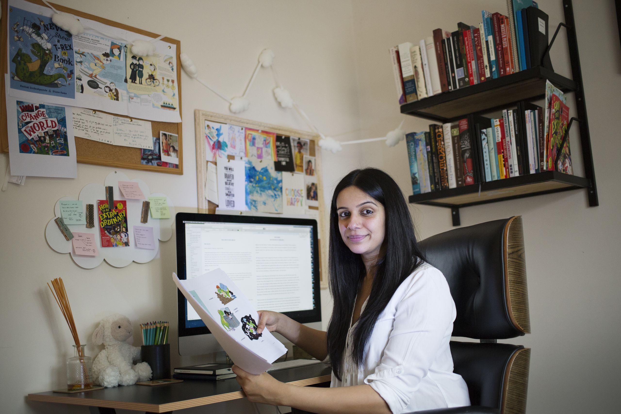 Rashmi at her desk, with books and pictures around her. Rashmi is holding some of the illustrations for her books.
